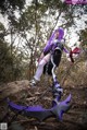 A woman with purple hair is sitting on a rock in the woods.